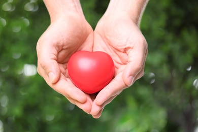 Young man holding red heart on blurred green background, closeup. Donation concept