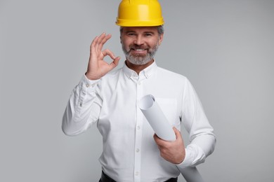 Photo of Architect in hard hat holding draft and showing ok gesture on grey background