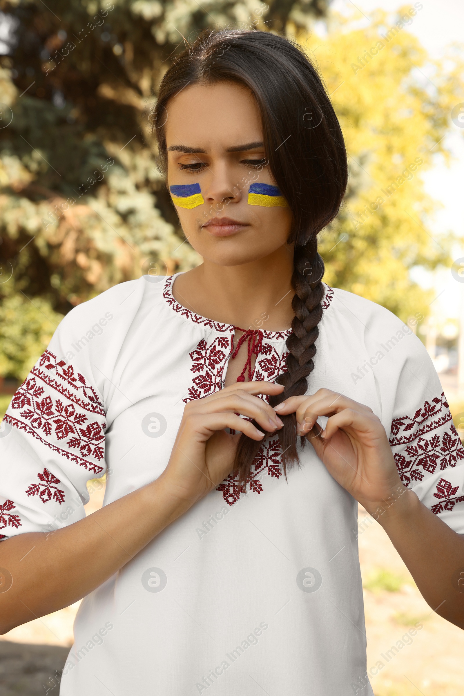 Photo of Sad young woman with drawings of Ukrainian flag on face in park