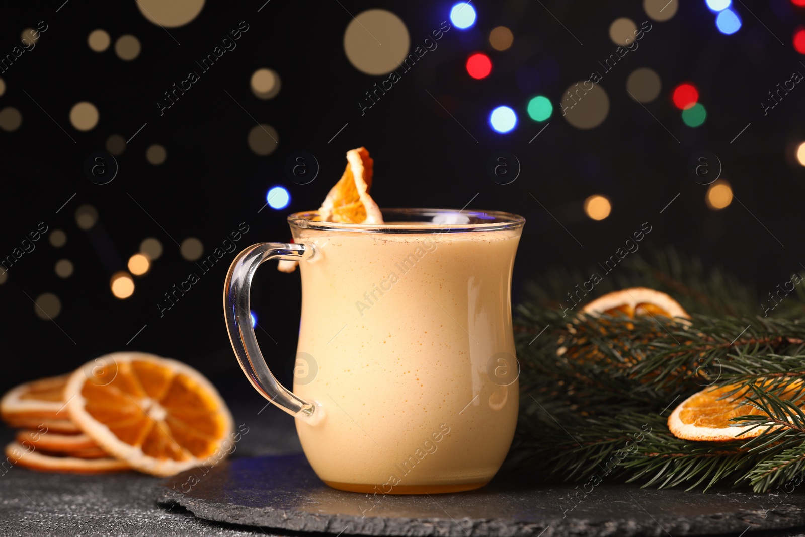 Photo of Glass cup of delicious eggnog with dried orange slice and fir branch on black table