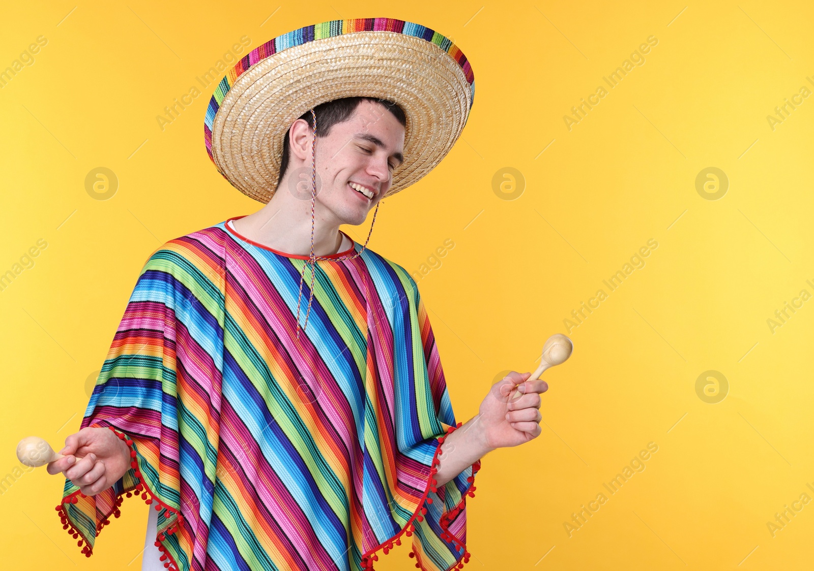 Photo of Young man in Mexican sombrero hat and poncho with maracas on yellow background. Space for text