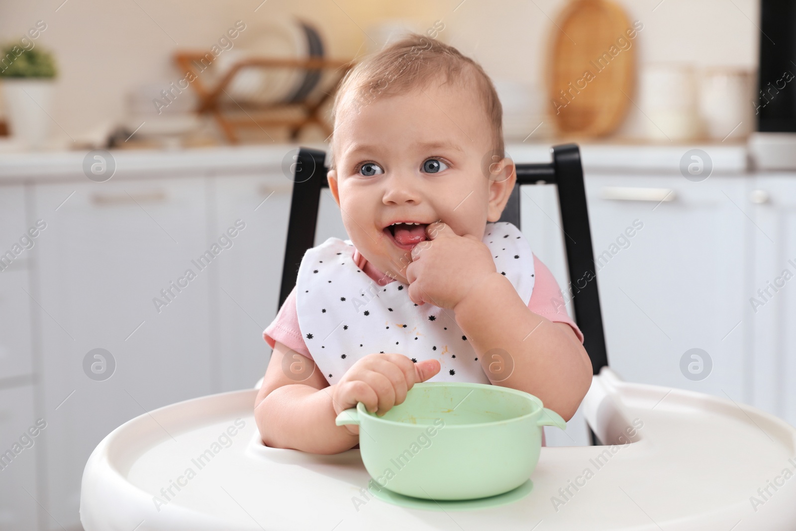 Photo of Cute little baby with bowl in high chair at kitchen