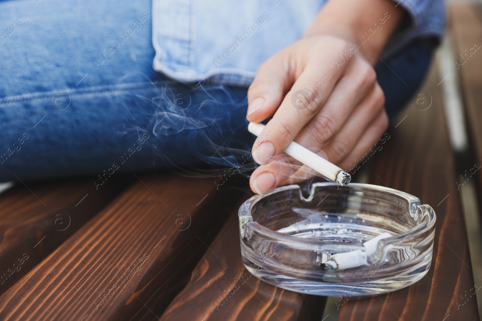 Photo of Woman holding cigarette over glass ashtray on bench outdoors, closeup