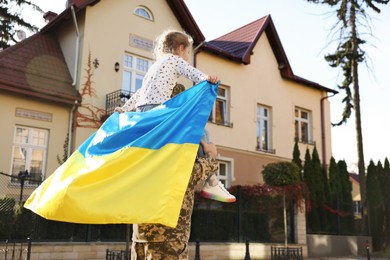 Soldier in military uniform carrying his daughter on shoulders and Ukrainian flag on city street, space for text. Family reunion