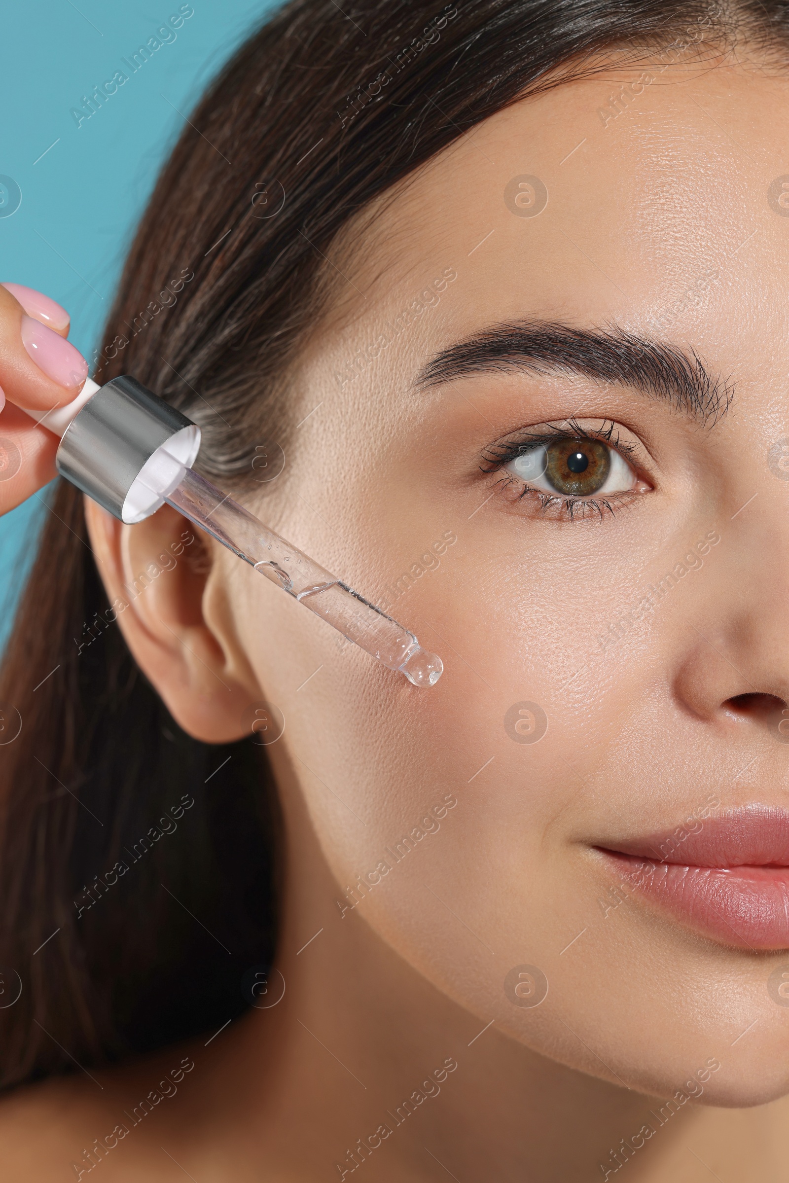 Photo of Beautiful young woman applying serum onto her face on light blue background, closeup