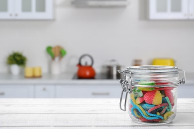 Glass jar with tasty gummy candies on white wooden table in kitchen. Space for text