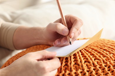 Photo of Man writing message in greeting card on orange
knitted pouf, closeup