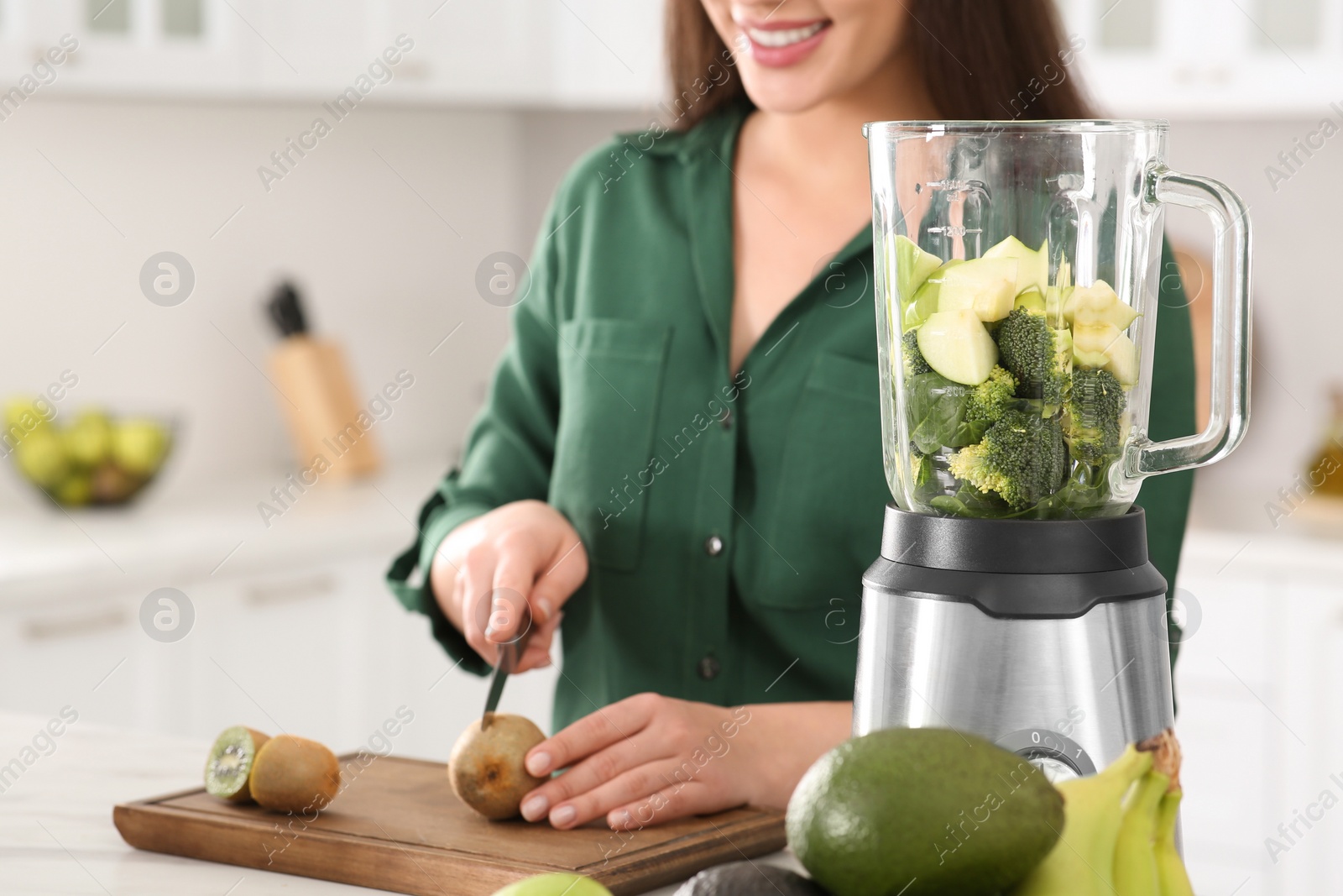 Photo of Young woman cutting kiwi for smoothie at white table in kitchen, closeup