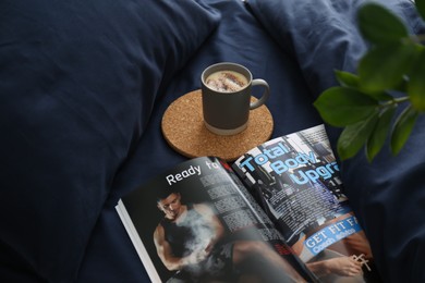 Photo of Open magazine and cup of coffee on bed with stylish silky linens, above view