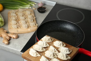 Photo of Wooden board with uncooked gyoza near frying pan on stove