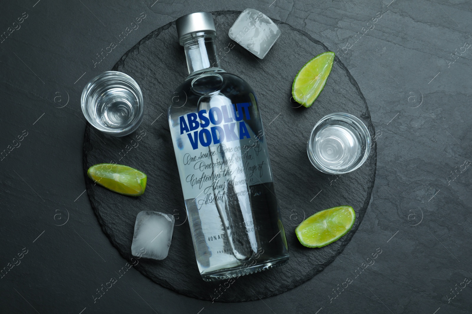Photo of MYKOLAIV, UKRAINE - OCTOBER 04, 2019: Absolut vodka, lime, ice cubes and shot glasses on black table, flat lay