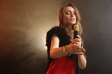 Beautiful young woman in stylish dress with microphone singing on dark background in color lights and smoke