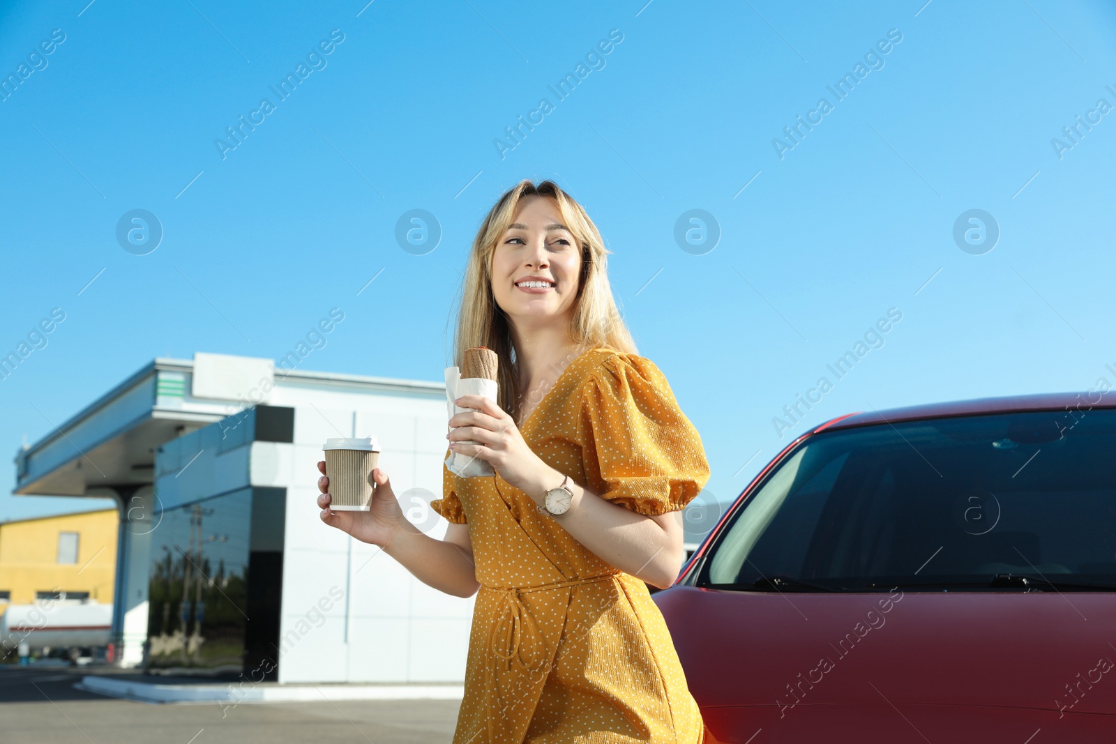 Photo of Beautiful young woman with coffee and hot dog near car at gas station