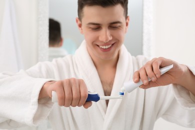 Photo of Man squeezing toothpaste from tube onto electric toothbrush in bathroom, selective focus