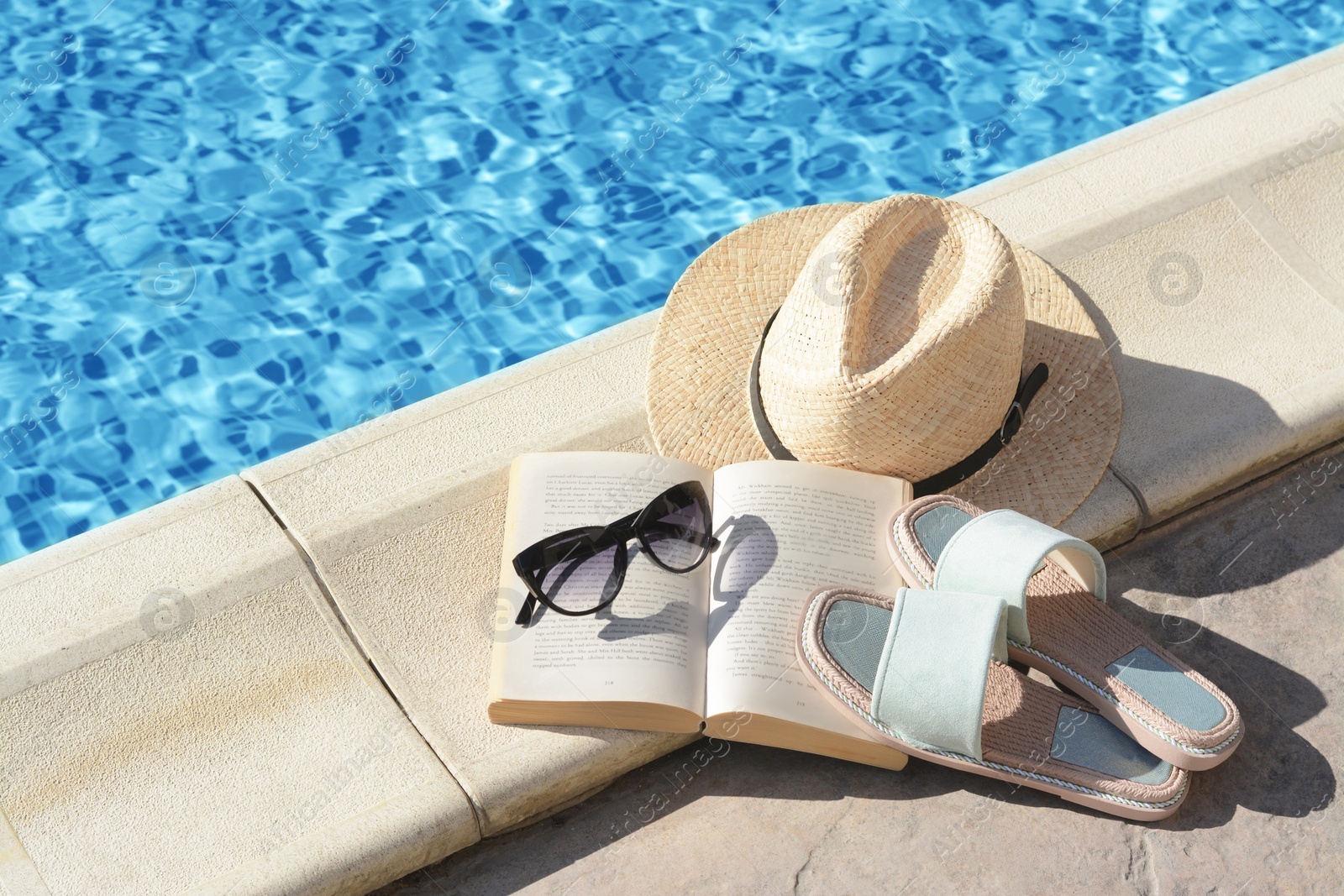 Photo of Stylish sunglasses, slippers, straw hat and book at poolside on sunny day. Beach accessories