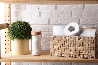 Toilet paper rolls in wicker basket, floral decor and cotton pads on wooden shelf against white brick wall