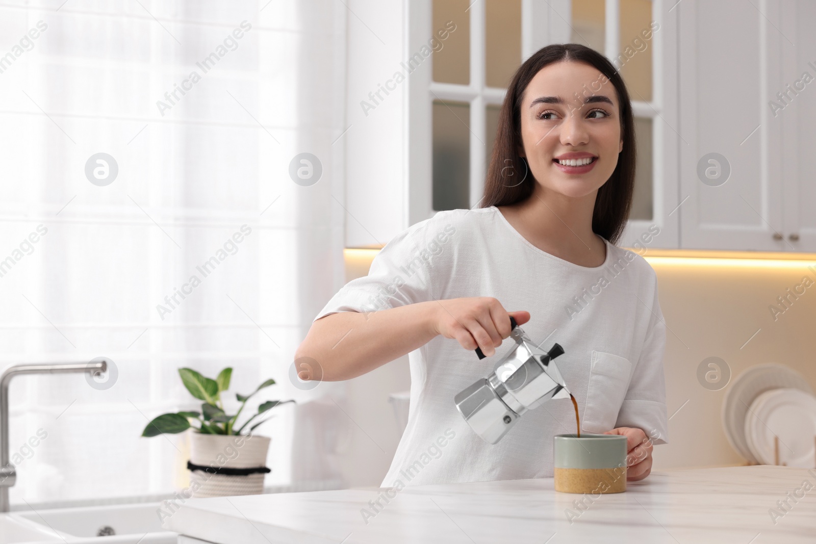 Photo of Happy woman pouring coffee from jezve into cup at white marble table in kitchen, space for text. Lazy morning