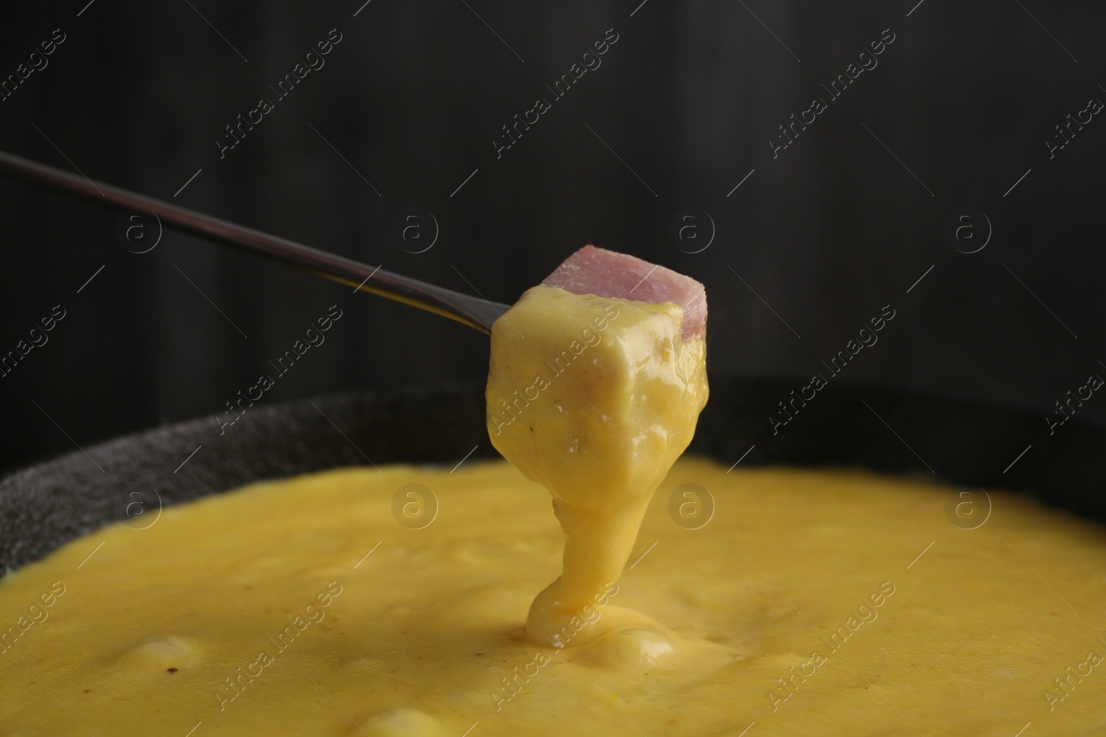 Photo of Dipping piece of ham into fondue pot with tasty melted cheese against dark gray background, closeup