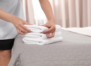 Young maid putting stack of fresh towels on bed in hotel room, closeup