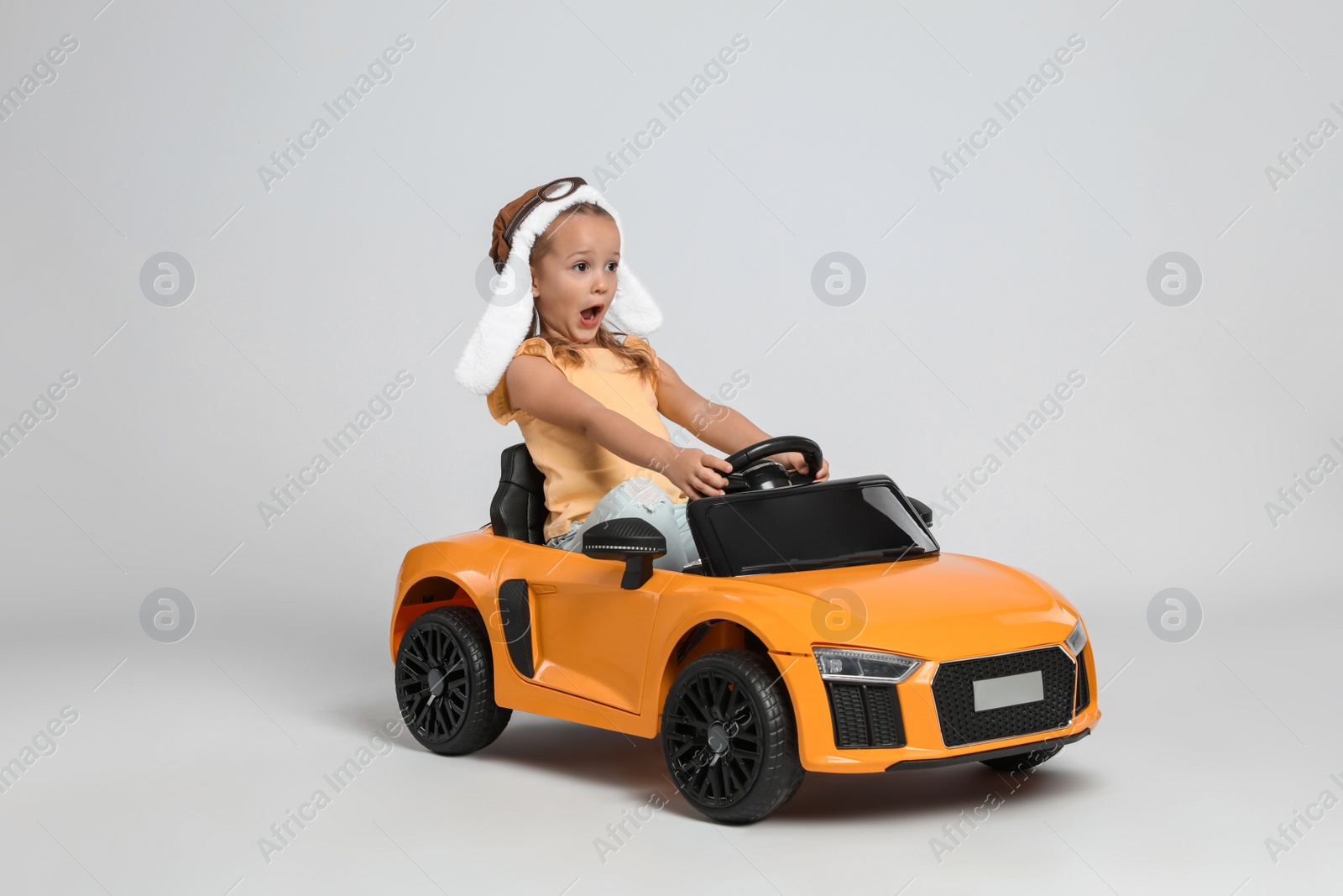 Photo of Cute little girl in pilot hat driving children's electric toy car on grey background