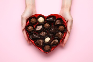 Woman holding heart shaped box with delicious chocolate candies on pink background, top view