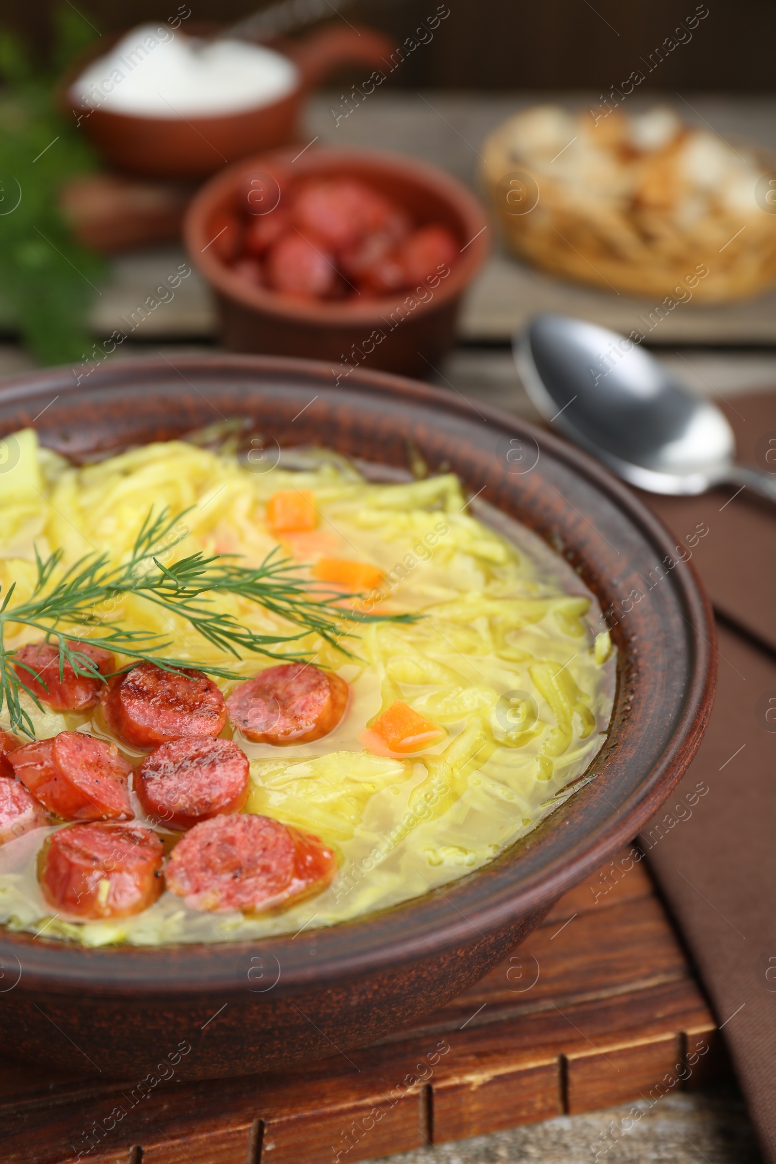 Photo of Bowl of delicious sauerkraut soup with smoked sausages and dill on wooden board, closeup