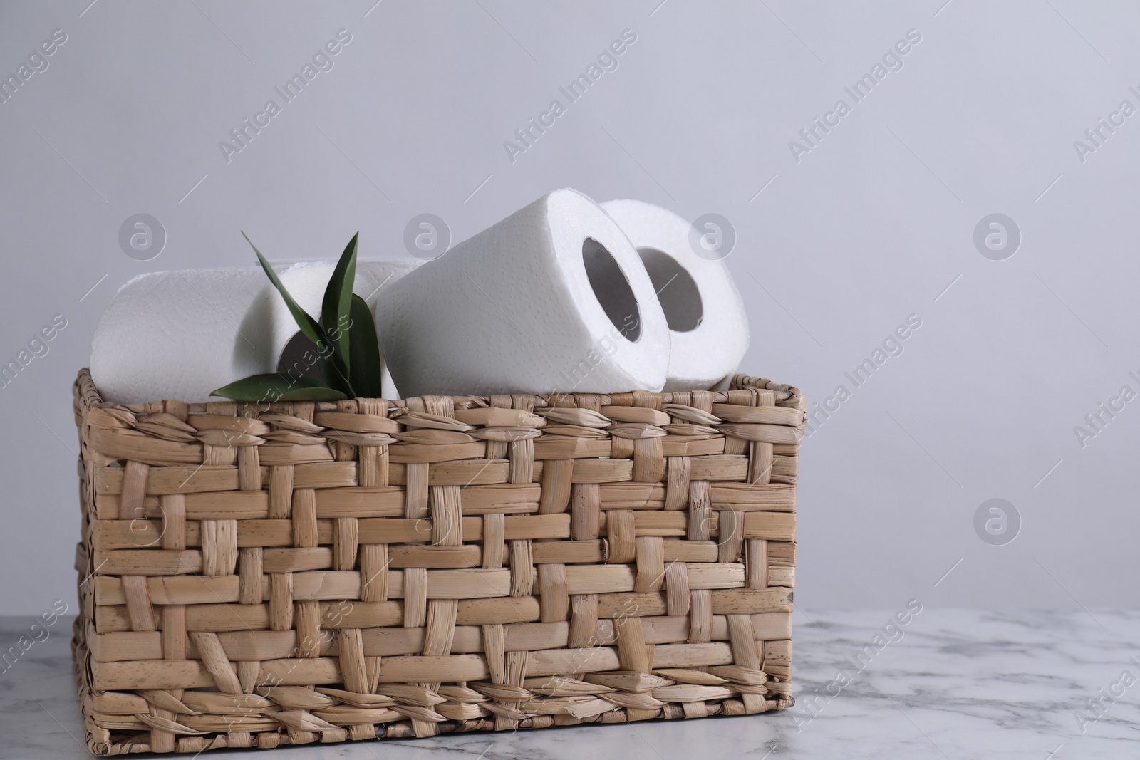 Photo of Toilet paper rolls and green leaves in wicker basket on white marble table, space for text