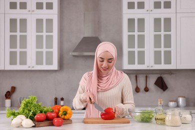 Muslim woman making delicious salad with vegetables at white table in kitchen