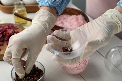 Photo of Woman in gloves filling bath bomb mold with flower buds at white table, closeup