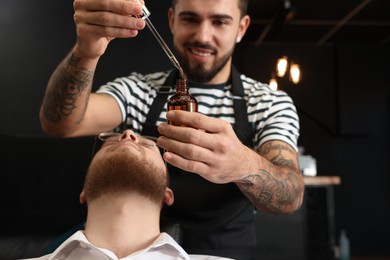 Hairdresser with beard oil near client in barbershop, closeup. Professional shaving service