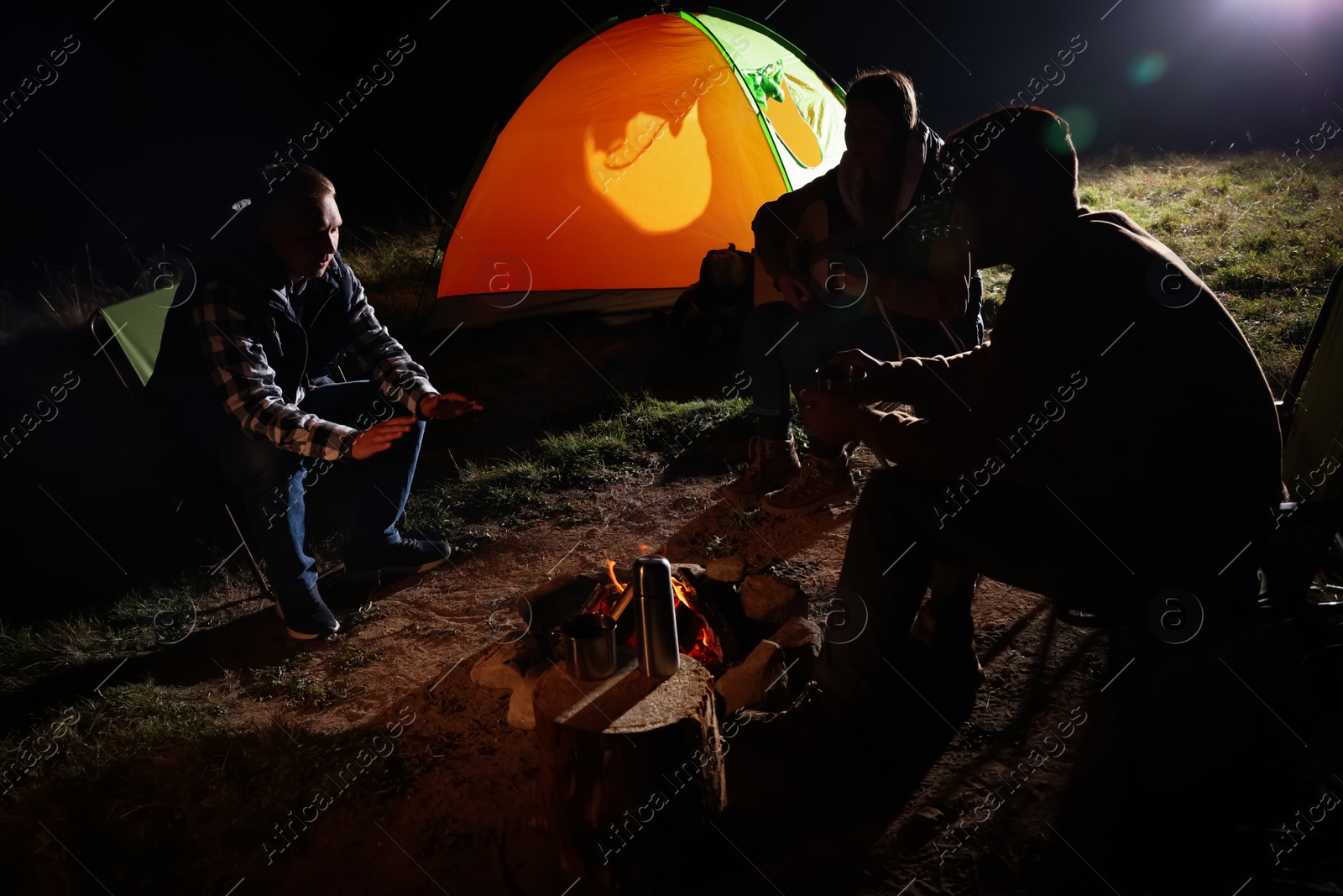 Photo of Group of friends with guitar near bonfire and camping tent outdoors at night