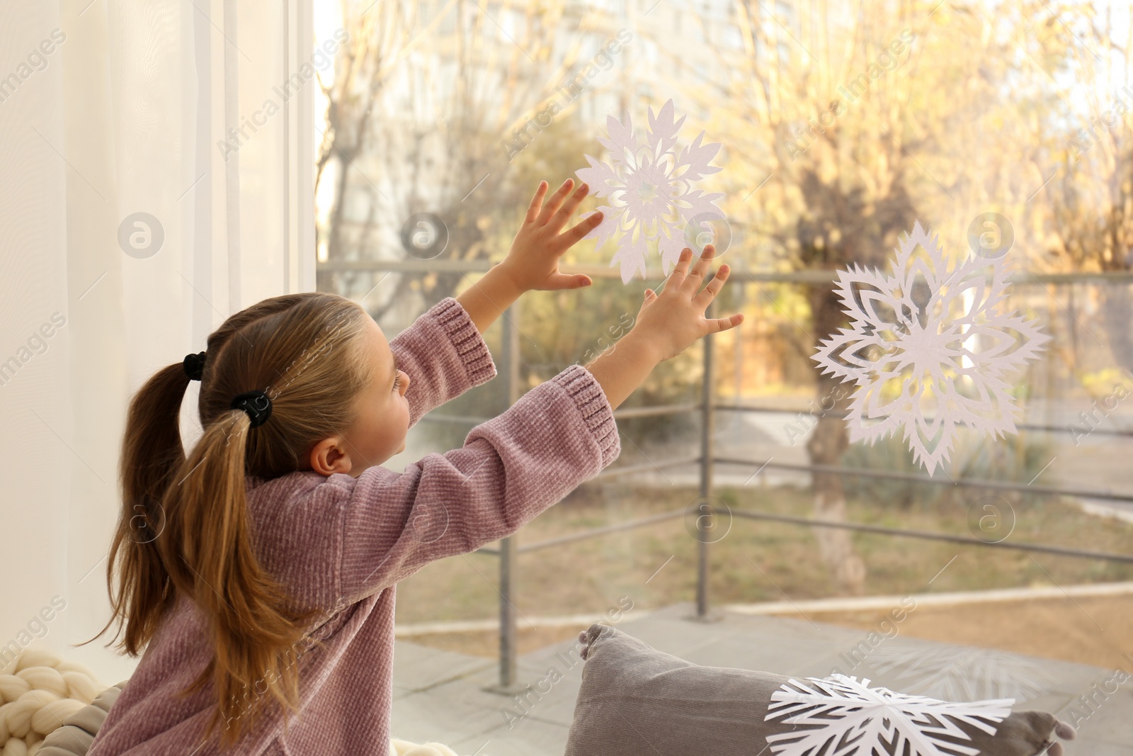 Photo of Little girl decorating window with paper snowflake indoors