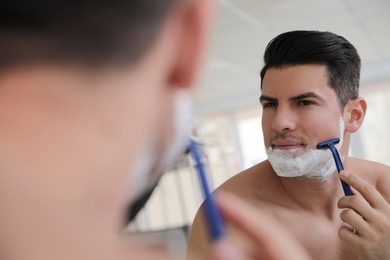 Handsome man shaving near mirror in bathroom