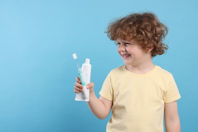 Cute little boy holding electric toothbrush and tube of toothpaste on light blue background, space for text