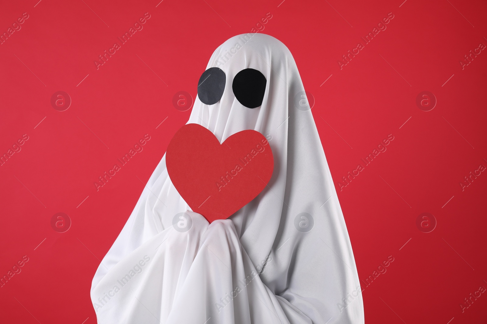 Photo of Cute ghost. Person covered with white sheet holding heart on red background