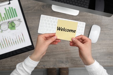 Image of Woman holding paper note with word Welcome over her office desk, top view