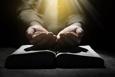 Image of Christian man with Bible holding hands under holy light in darkness, closeup
