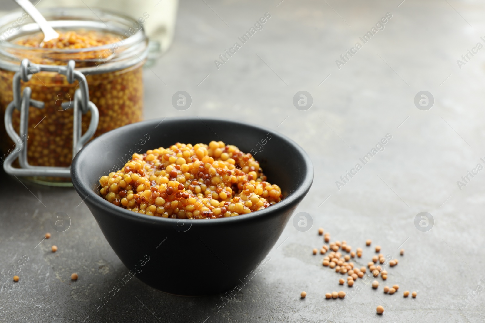 Photo of Whole grain mustard in bowl and dry seeds on grey table. Space for text