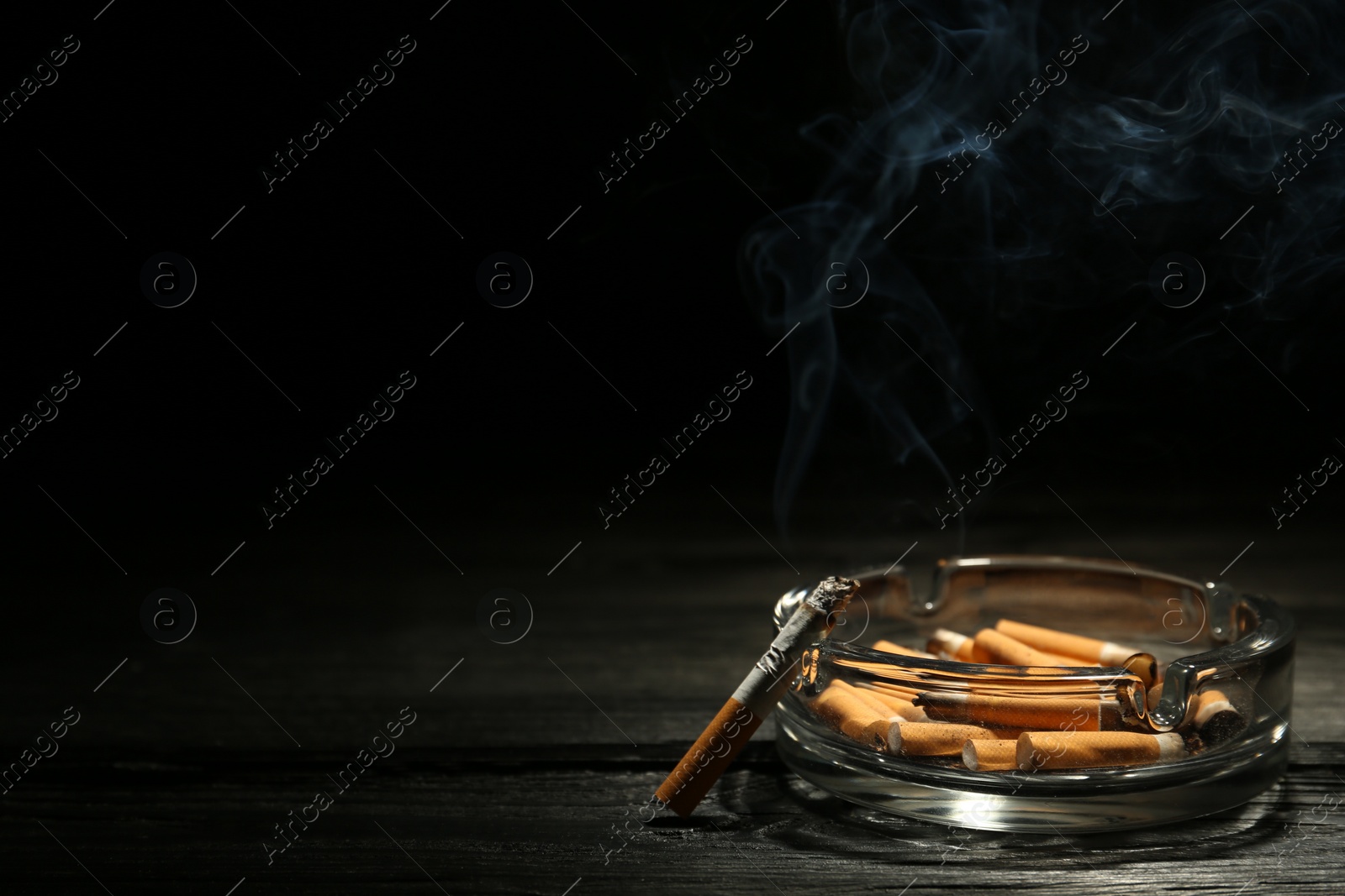 Photo of Glass ashtray with stubs and smoldering cigarette on dark wooden table against black background. Space for text