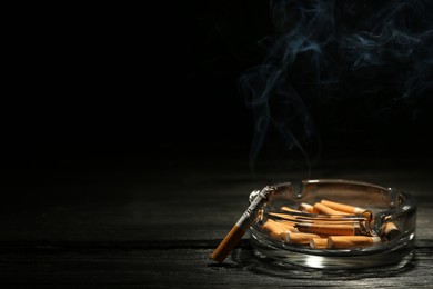 Photo of Glass ashtray with stubs and smoldering cigarette on dark wooden table against black background. Space for text