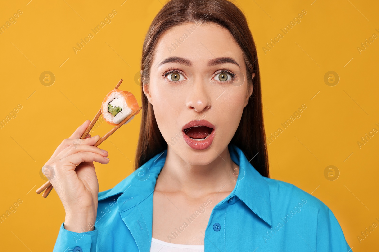 Photo of Emotional young woman holding sushi roll with chopsticks on orange background