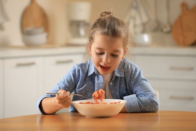 Photo of Cute little girl eating tasty pasta at table in kitchen