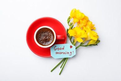 Photo of Cup of aromatic coffee, beautiful yellow freesias and Good Morning note on white background, flat lay