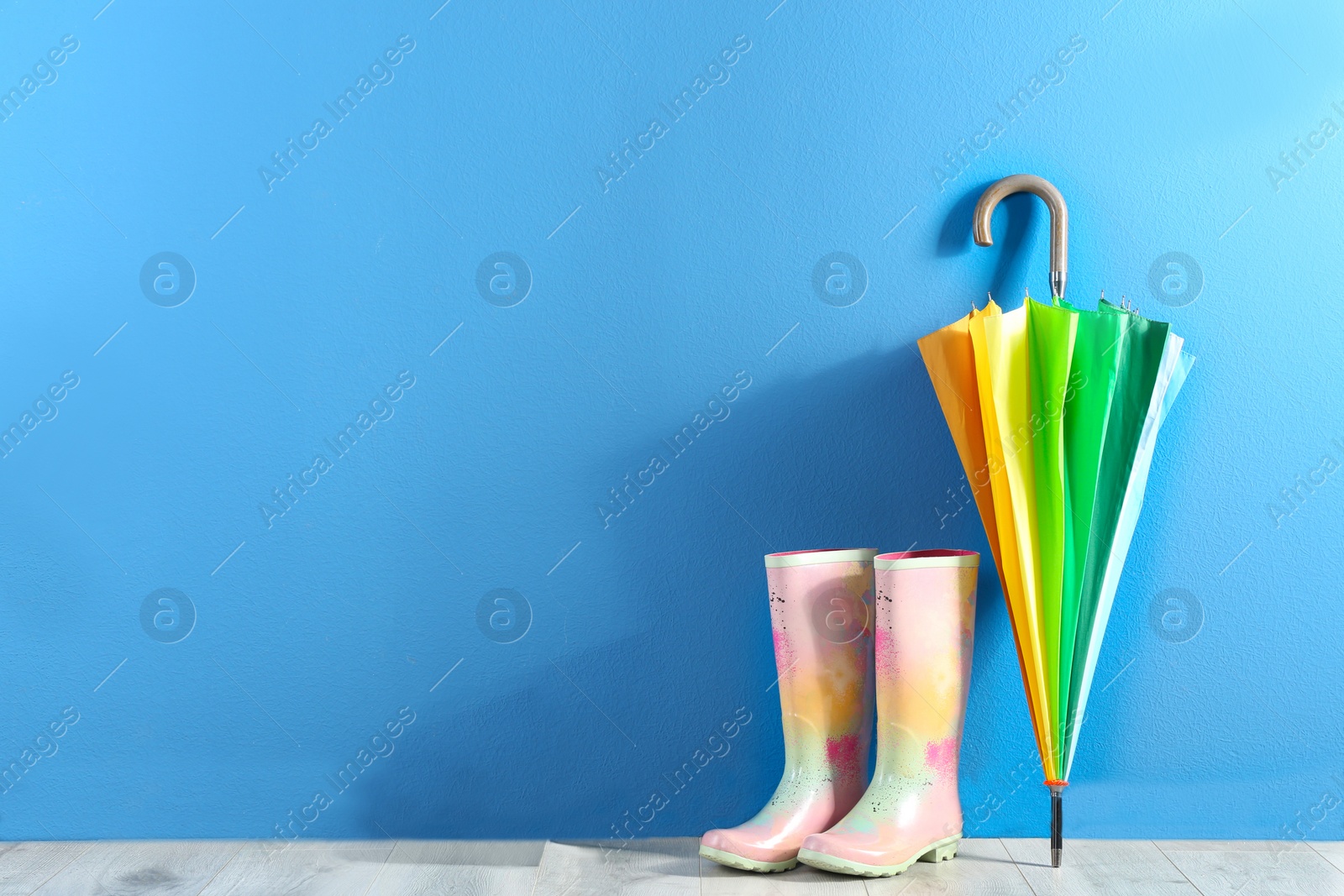 Photo of Beautiful colorful umbrella and rubber boots near blue wall. Space for text