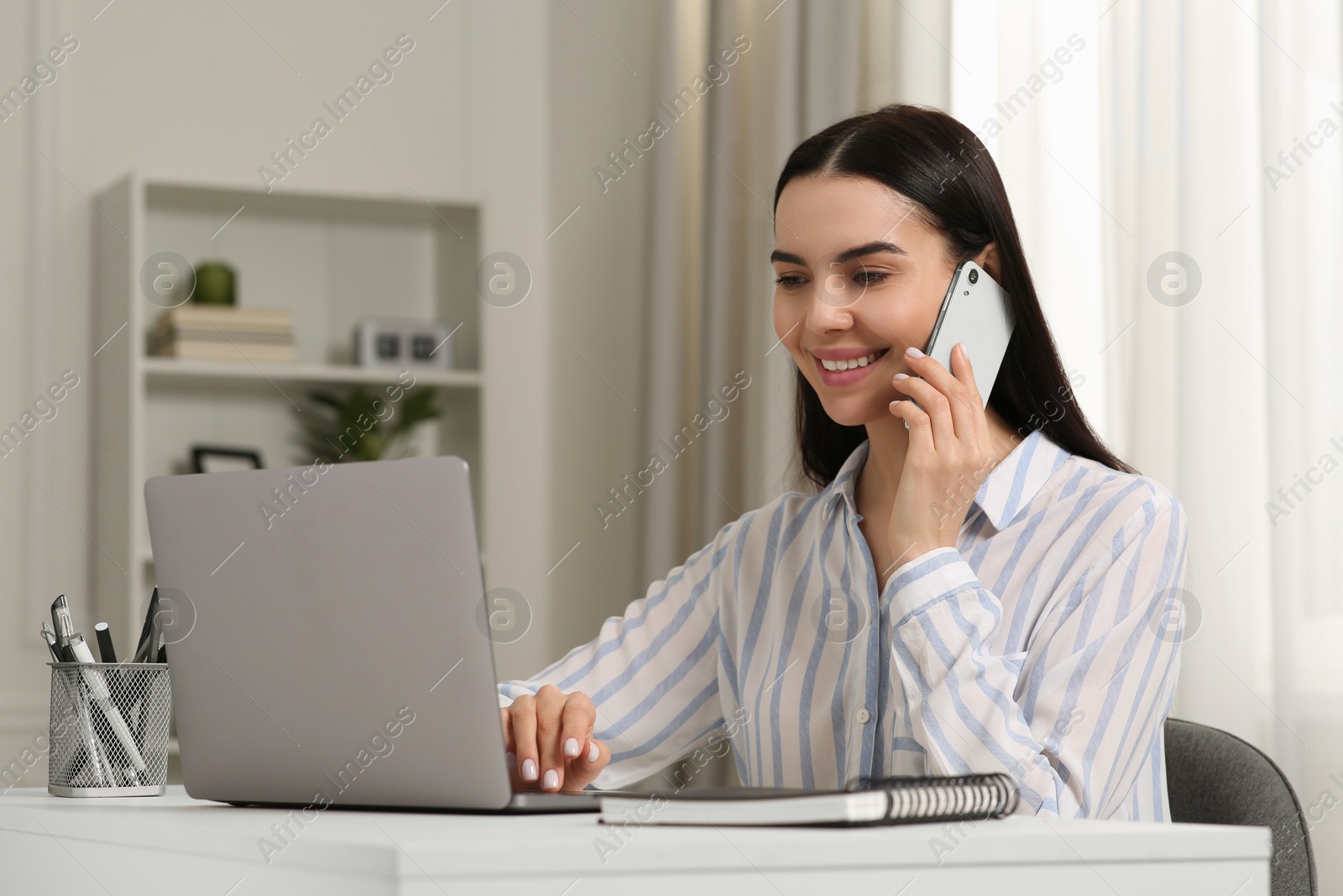 Photo of Happy woman talking on smartphone while working with laptop at white desk in room