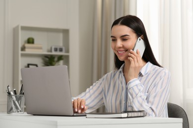 Photo of Happy woman talking on smartphone while working with laptop at white desk in room