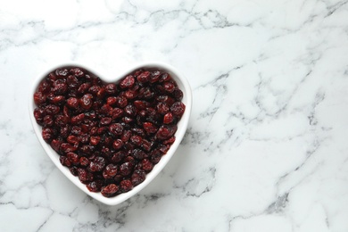 Photo of Heart shaped bowl with cranberries on marble table, top view with space for text. Dried fruit as healthy snack