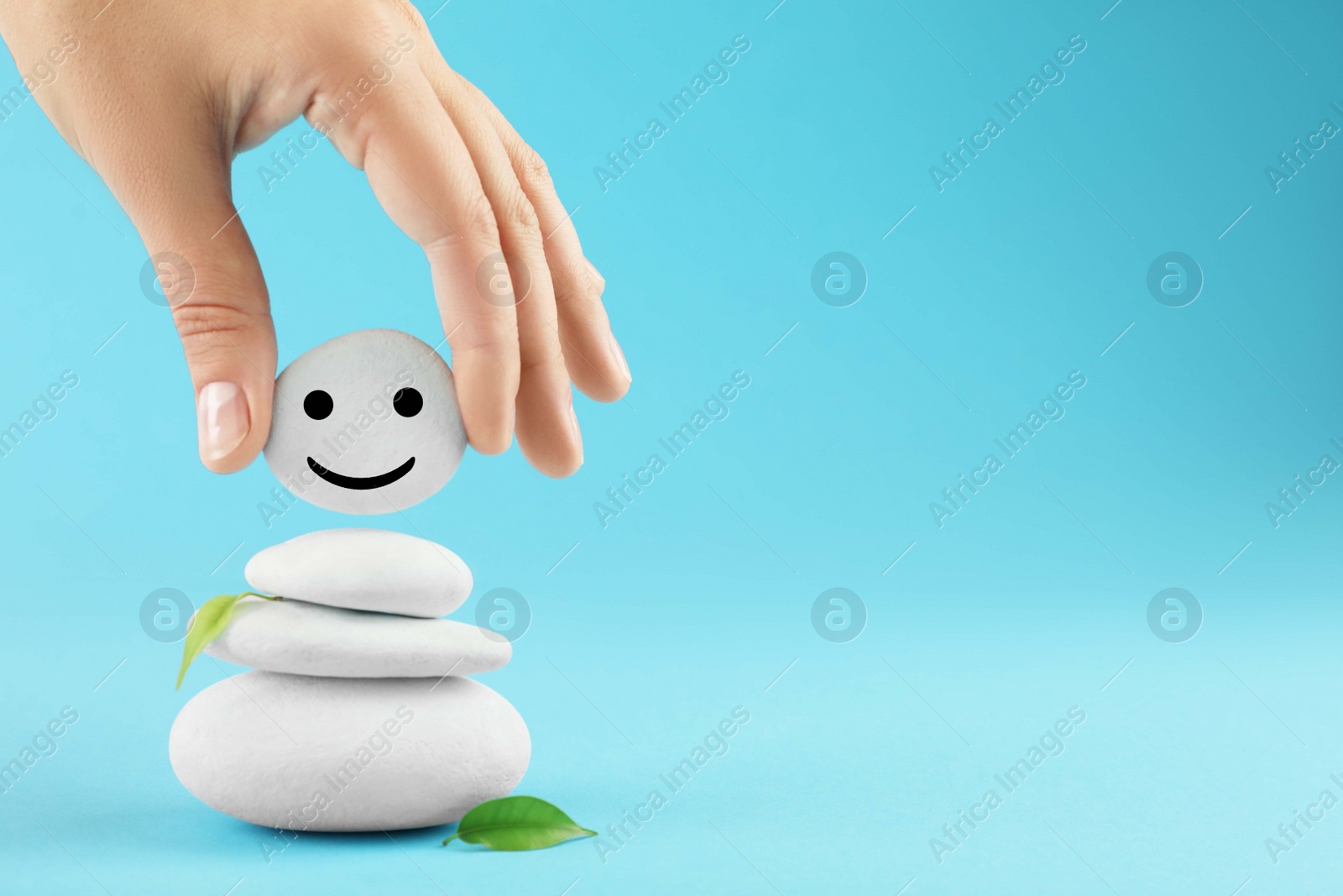 Photo of Woman putting stone with drawn happy face on stack against light blue background, closeup and space for text. Zen concept