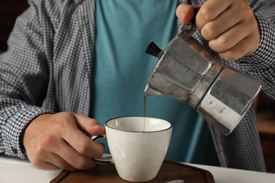 Man pouring aromatic coffee from moka pot into cup at white table, closeup