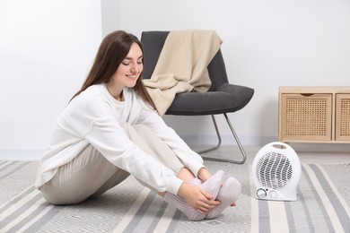 Photo of Young woman warming feet near electric fan heater at home